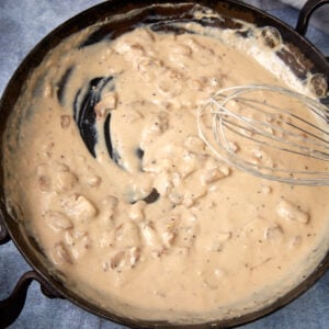 A skillet filled with creamy, light brown gravy containing small chunks, with a metal whisk resting in the sauce and a streak showing where the sauce has been stirred.