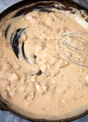 A skillet filled with creamy, light brown gravy containing small chunks, with a metal whisk resting in the sauce and a streak showing where the sauce has been stirred.