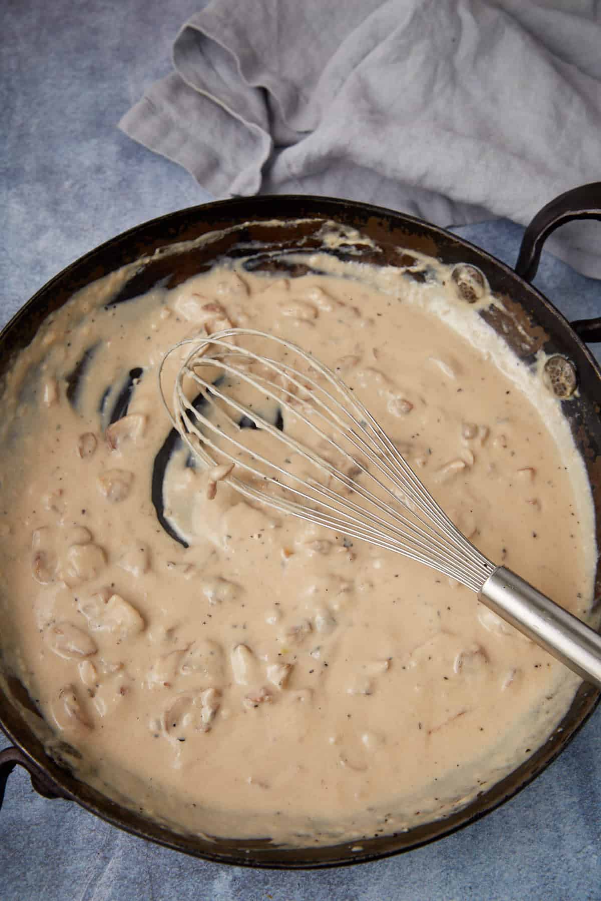A metal whisk rests in a skillet filled with creamy white gravy containing small chunks, set on a blue-gray surface with a light gray cloth nearby.