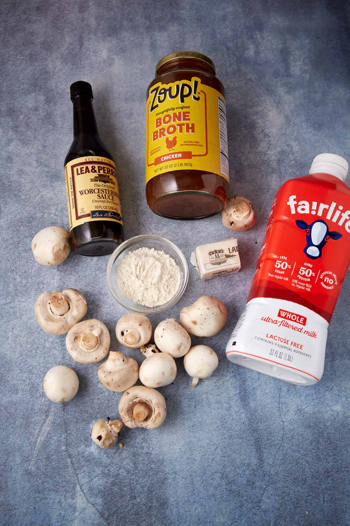 A flat lay of cooking ingredients including mushrooms, a bottle of Worcestershire sauce, chicken bone broth, a stick of butter, a bowl of flour, and a carton of lactose-free whole milk on a gray surface.