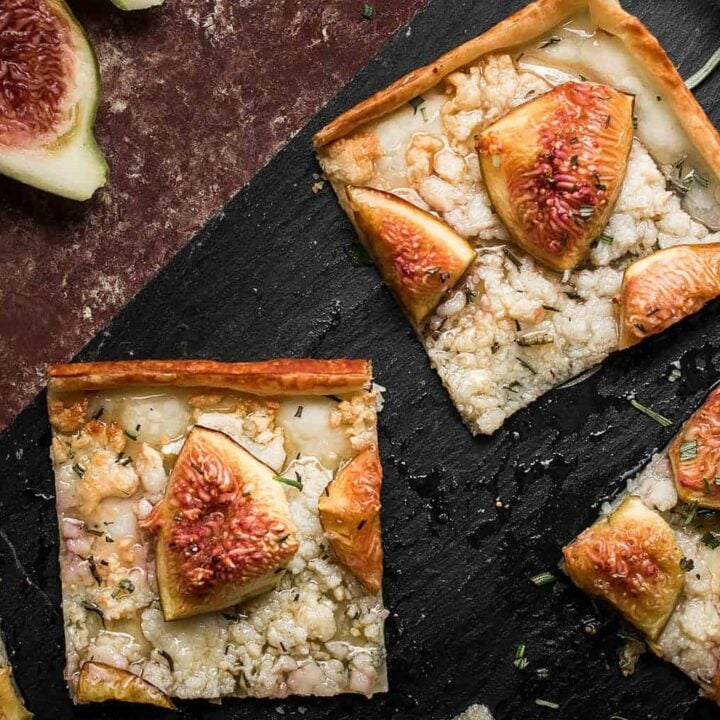 Three square slices of Goat Cheese and Fig Tart rest on a dark slate board, garnished with rosemary. Fresh figs, some whole and some halved, are placed nearby on a rustic surface.