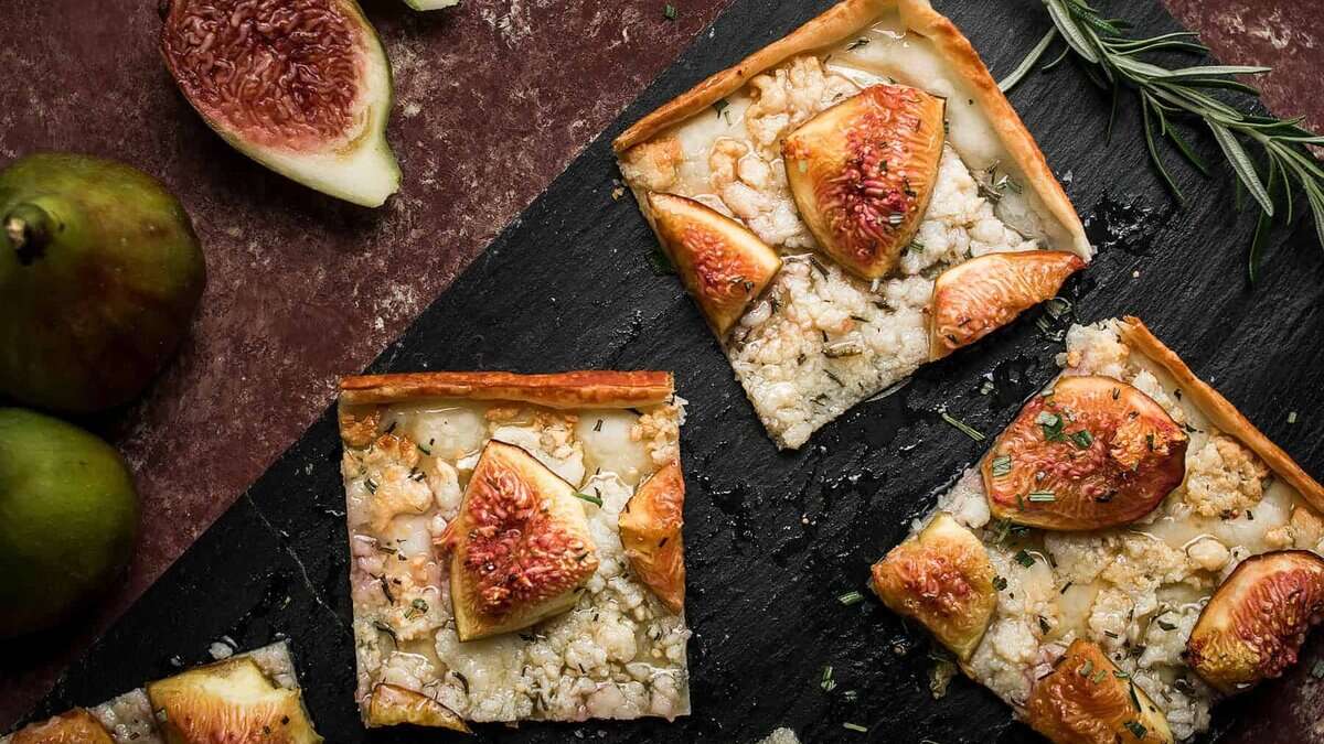 Three square slices of Goat Cheese and Fig Tart rest on a dark slate board, garnished with rosemary. Fresh figs, some whole and some halved, are placed nearby on a rustic surface.