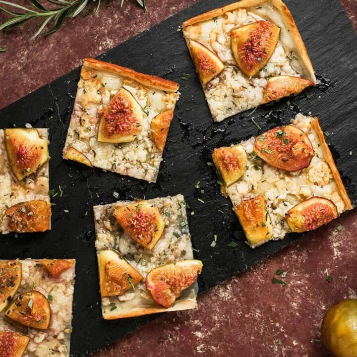 Five rectangular slices of Goat Cheese and Fig Tart topped with herbs are arranged on a black slate board. Whole fresh figs and sprigs of rosemary are scattered around on a rustic red surface.