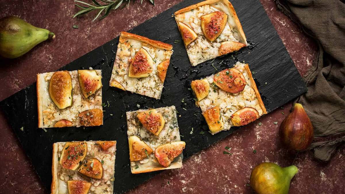 Five rectangular slices of Goat Cheese and Fig Tart topped with herbs are arranged on a black slate board. Whole fresh figs and sprigs of rosemary are scattered around on a rustic red surface.