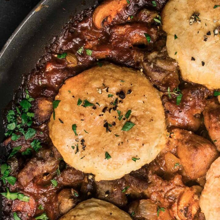 A skillet filled with a rich beef and Guinness pie stew of meat and vegetables, topped with golden, flaky biscuits and garnished with chopped parsley. A gray cloth napkin is partially visible in the corner.