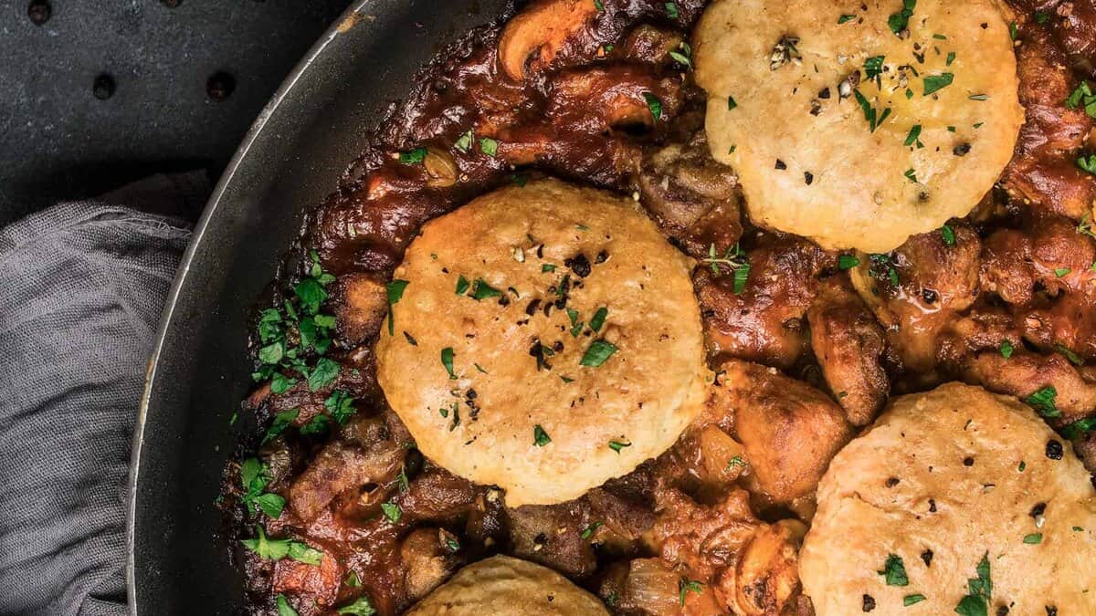 A skillet filled with a rich beef and Guinness pie stew of meat and vegetables, topped with golden, flaky biscuits and garnished with chopped parsley. A gray cloth napkin is partially visible in the corner.