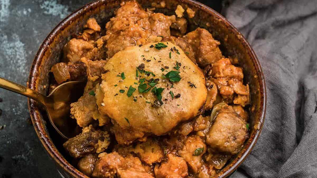 A bowl of hearty beef and Guinness pie stew with chunks of meat and vegetables, topped with a golden biscuit and garnished with chopped herbs. In the background, a pan with more stew and biscuits is visible.