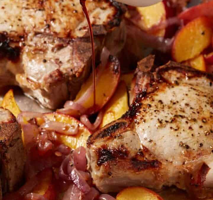Pork chops and peaches on a sheet pan with balsamic vinegar being poured over the top.