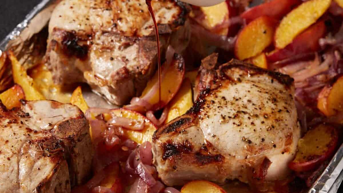 Pork chops and peaches on a sheet pan with balsamic vinegar being poured over the top. 