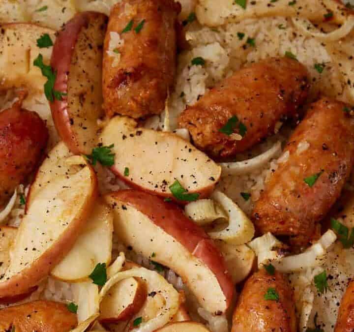 Sausage sheet pan dinner featuring sliced sausages, apple wedges, and onions baked on a foil-lined tray, garnished with chopped parsley and cracked black pepper.