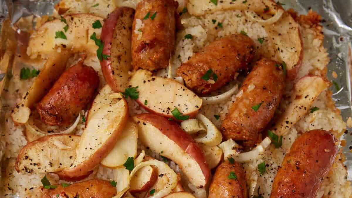 Sausage sheet pan dinner featuring sliced sausages, apple wedges, and onions baked on a foil-lined tray, garnished with chopped parsley and cracked black pepper.