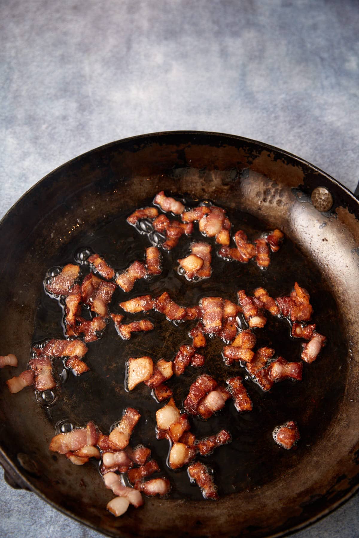 Crispy bacon pieces sizzling in a dark, well-used frying pan, with bacon fat visible in the pan, set against a light, softly textured background.