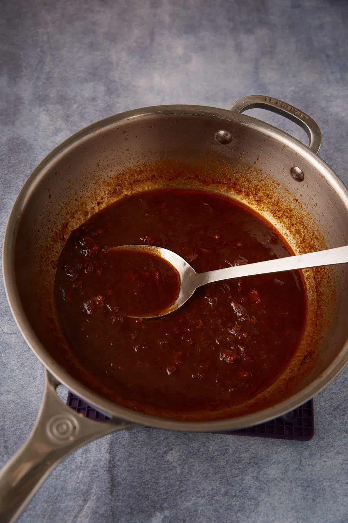 A stainless steel pan with a reddish-brown sauce and chunks sits on a blue surface. A metal spoon rests inside the pan, partially submerged in the sauce.