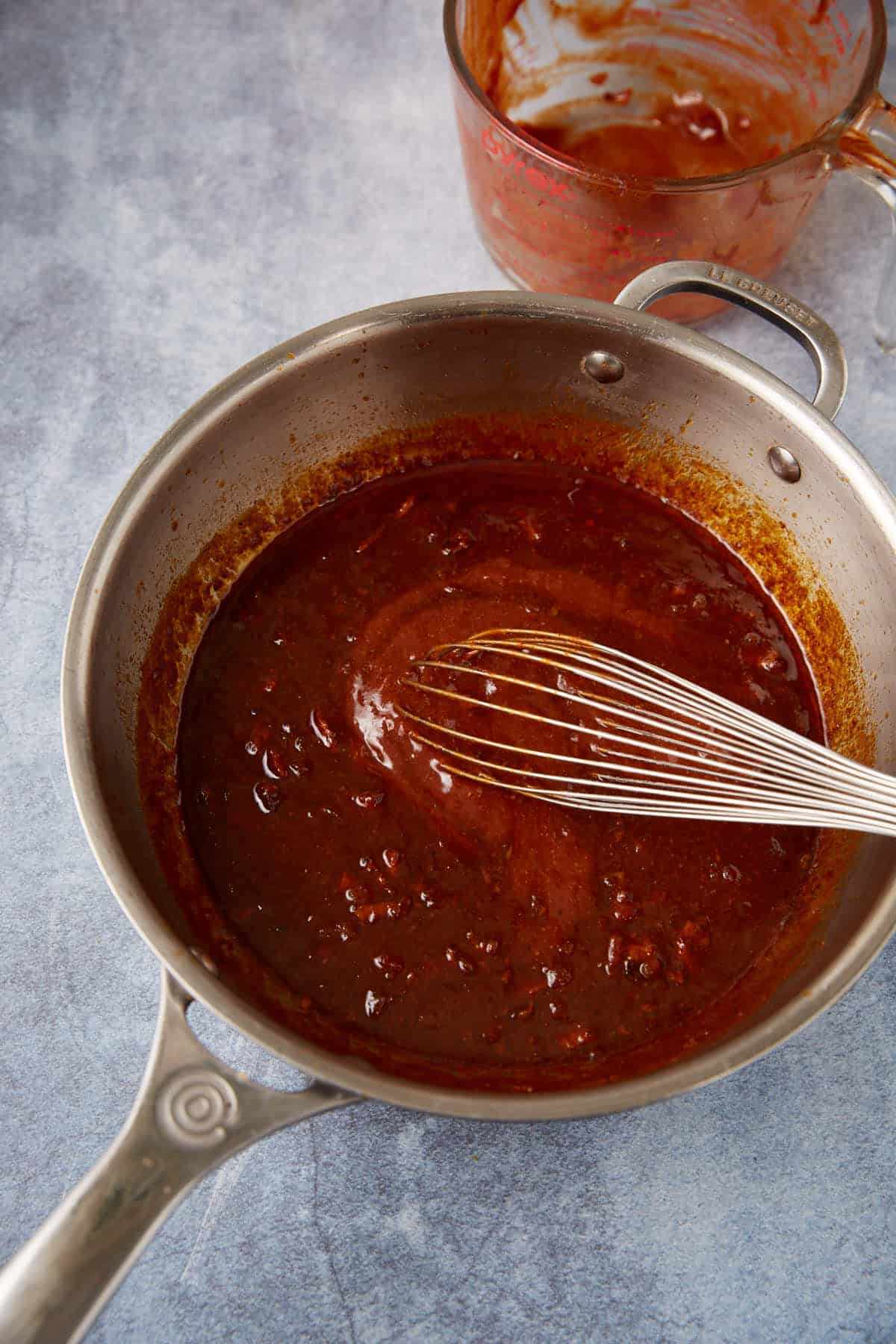 A saucepan filled with a thick, dark reddish-brown sauce being whisked; in the background, a measuring cup with sauce residue sits on a blue textured surface.
