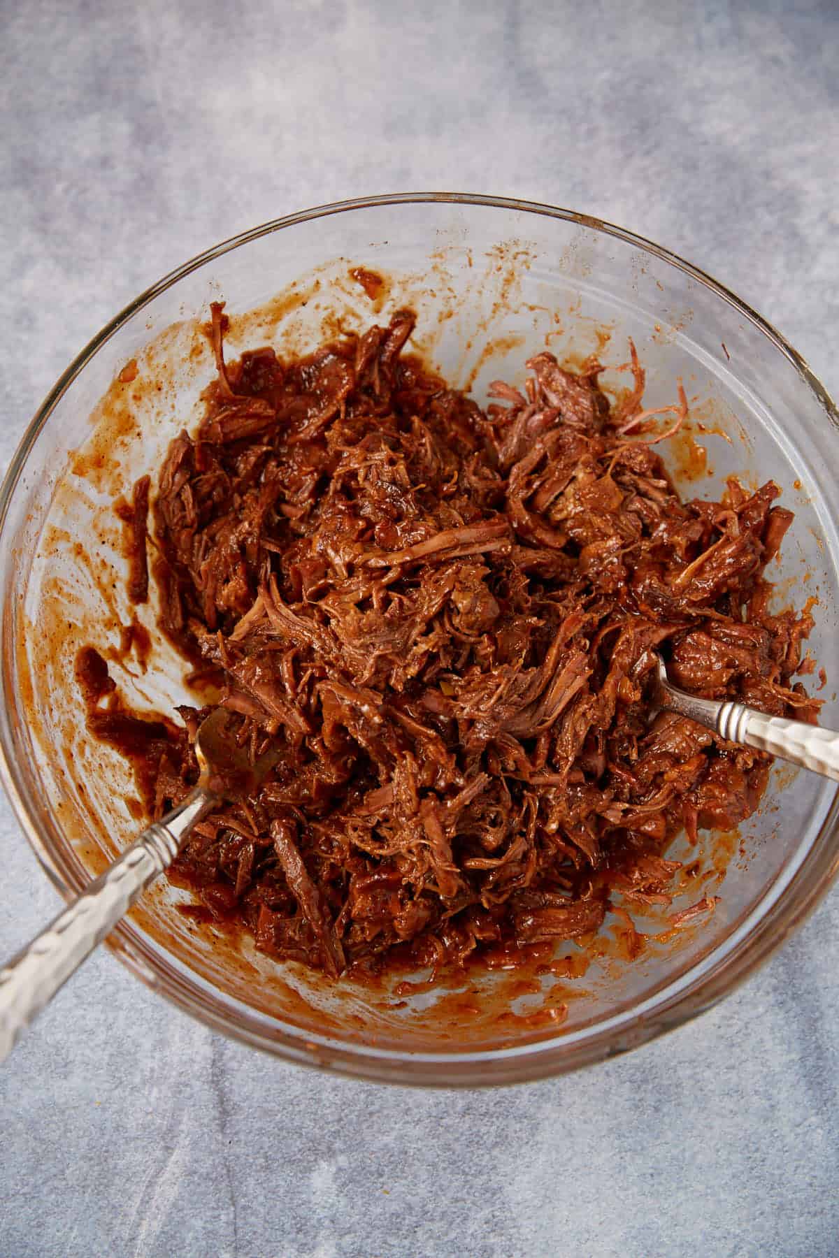 A glass bowl filled with shredded, saucy beef, mixed with two forks. The beef appears tender and coated in a rich, reddish-brown sauce. The bowl is set on a light gray surface.