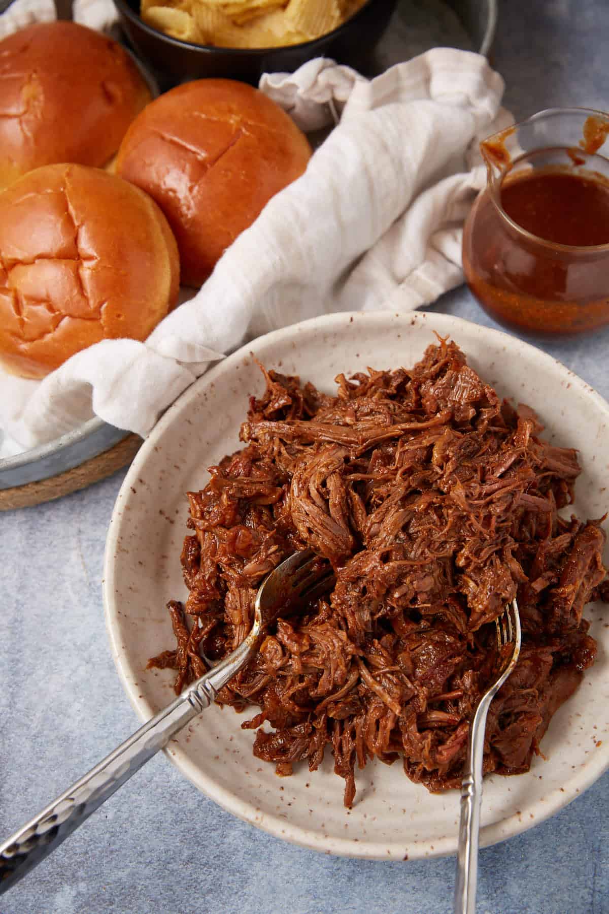 A plate of shredded barbecue beef with two forks, next to a jar of barbecue sauce, brioche buns, and a basket of potato chips on a light blue surface.