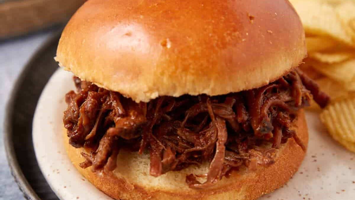 A close-up of a shredded beef sandwich on a round bun, served on a plate next to a small pile of potato chips. The background shows part of a bun and a blurred napkin.