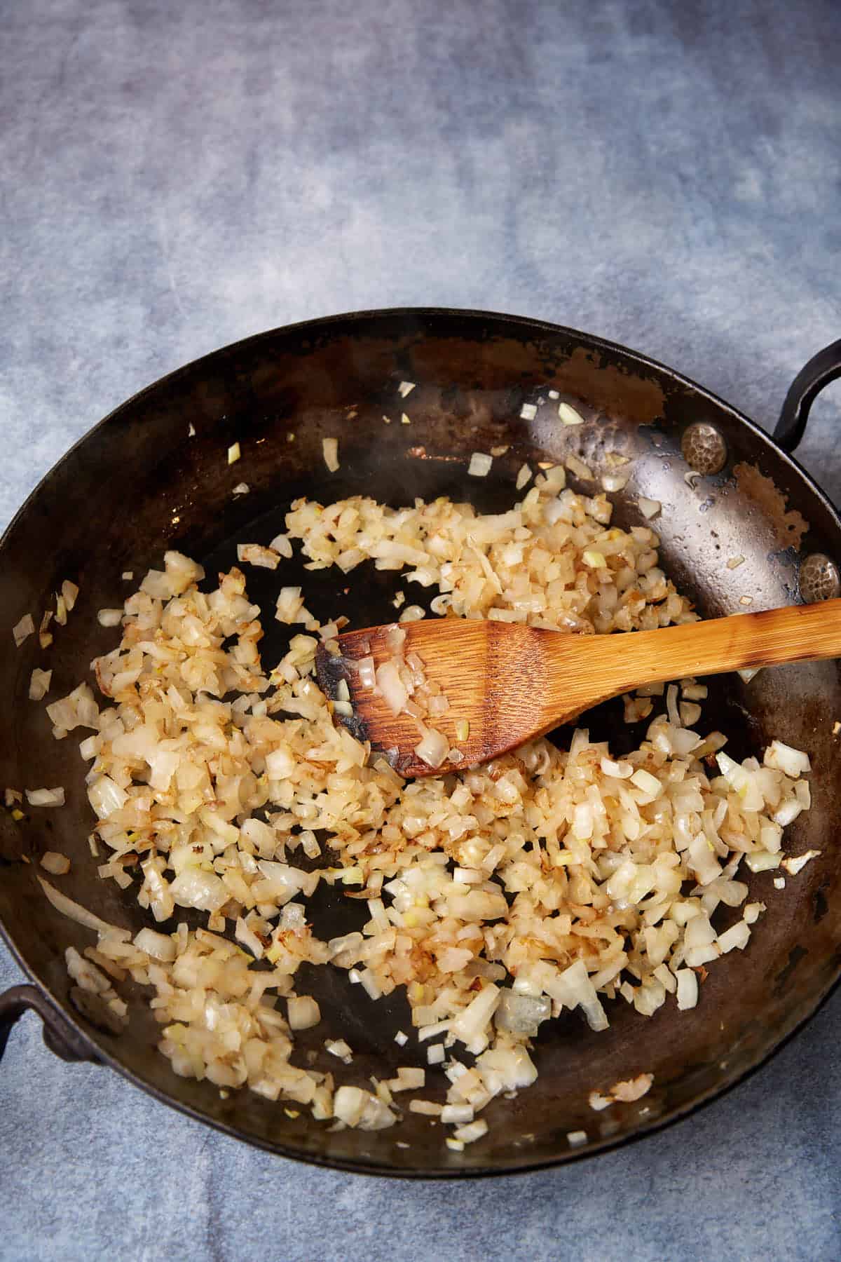 Chopped onions being sautéed and lightly browned in a large, dark frying pan with a wooden spatula resting on top.