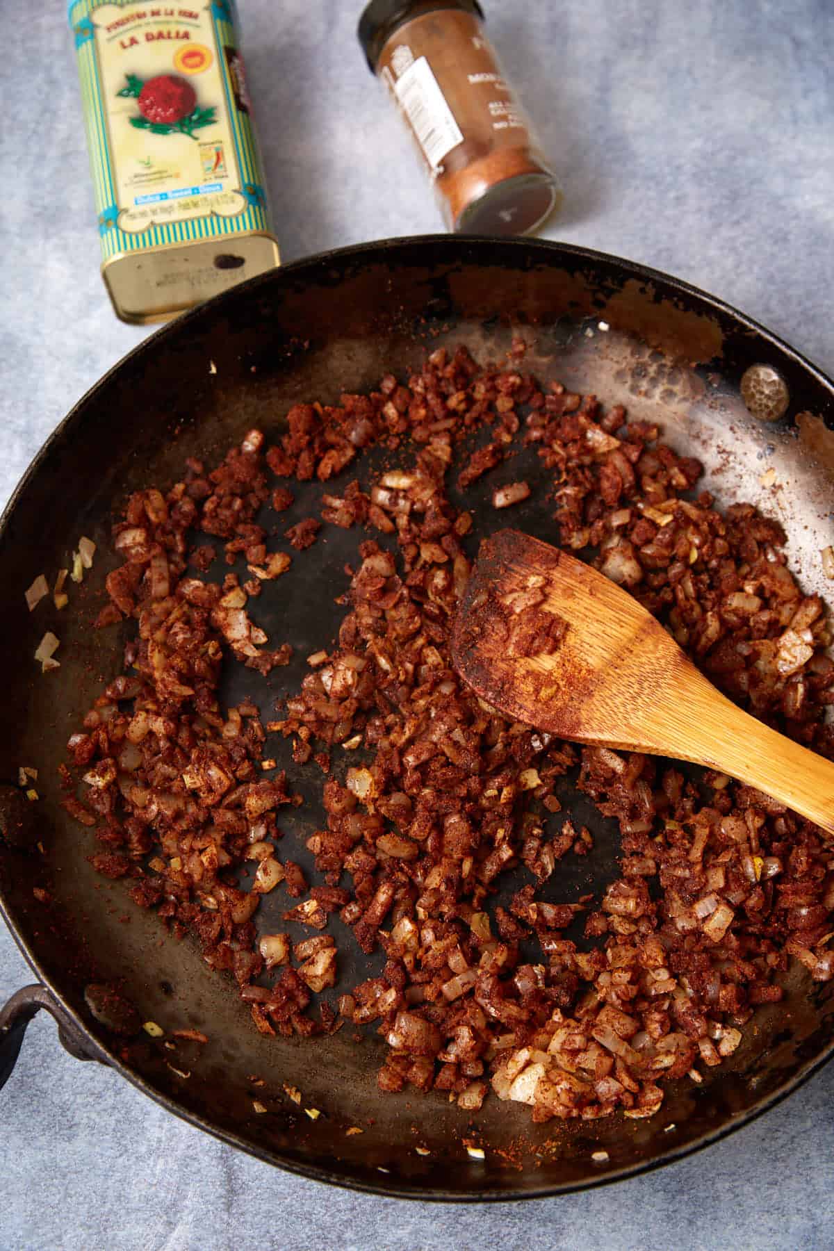 A skillet with finely chopped onions and chorizo being sautéed, stirred with a wooden spoon. Nearby are a tin of smoked paprika and a small jar of spice on a light surface.