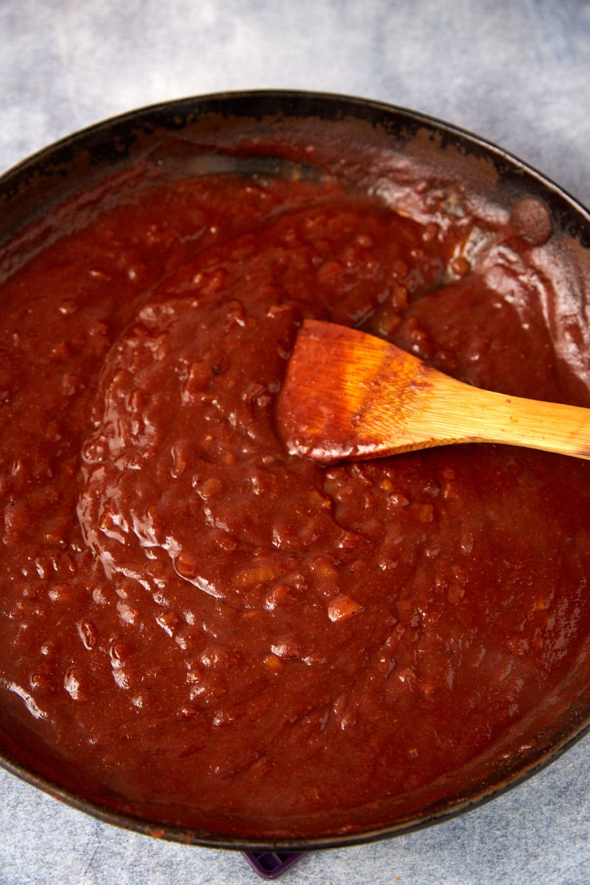 A close-up of a pan filled with thick, chunky tomato sauce being stirred with a wooden spatula, on a blue textured background.