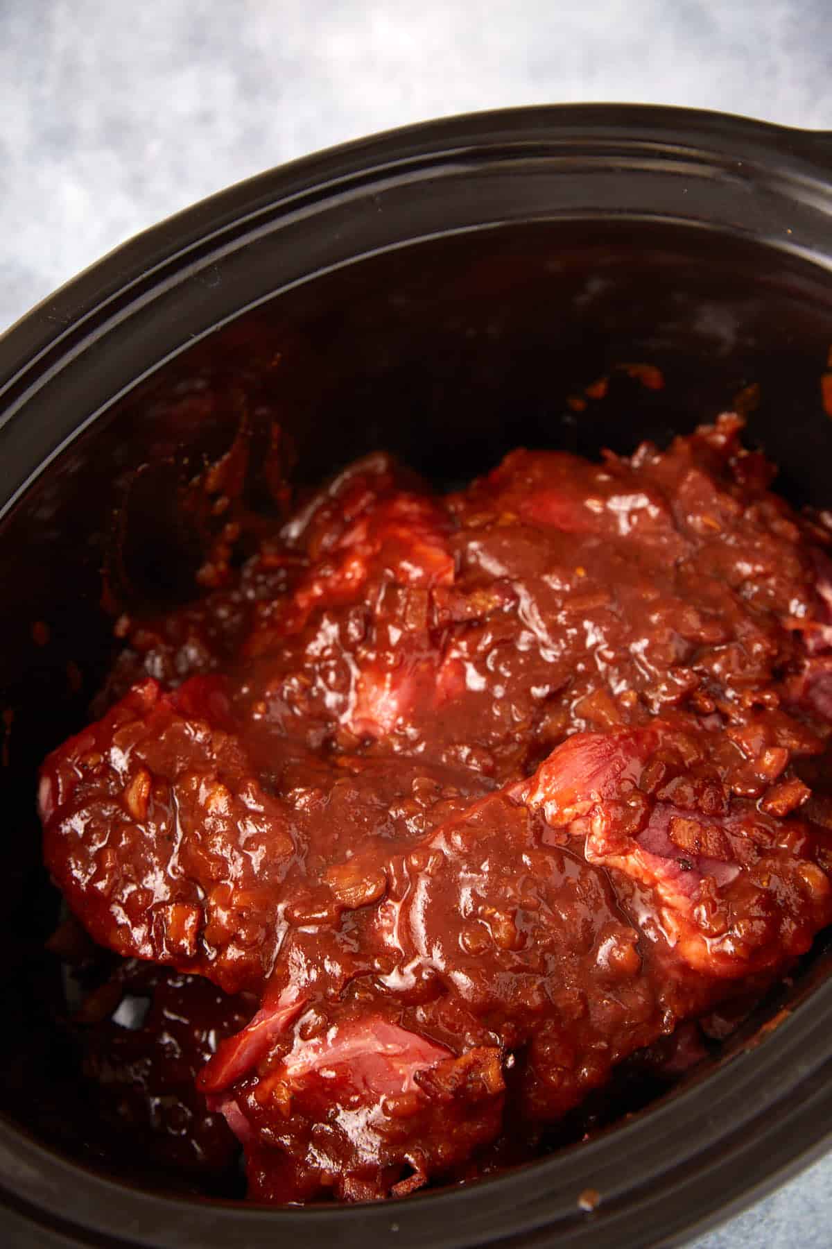 Raw meat covered in a thick, reddish-brown sauce sits inside a black slow cooker, ready to be cooked. The sauce appears chunky and rich, likely containing tomatoes and spices.