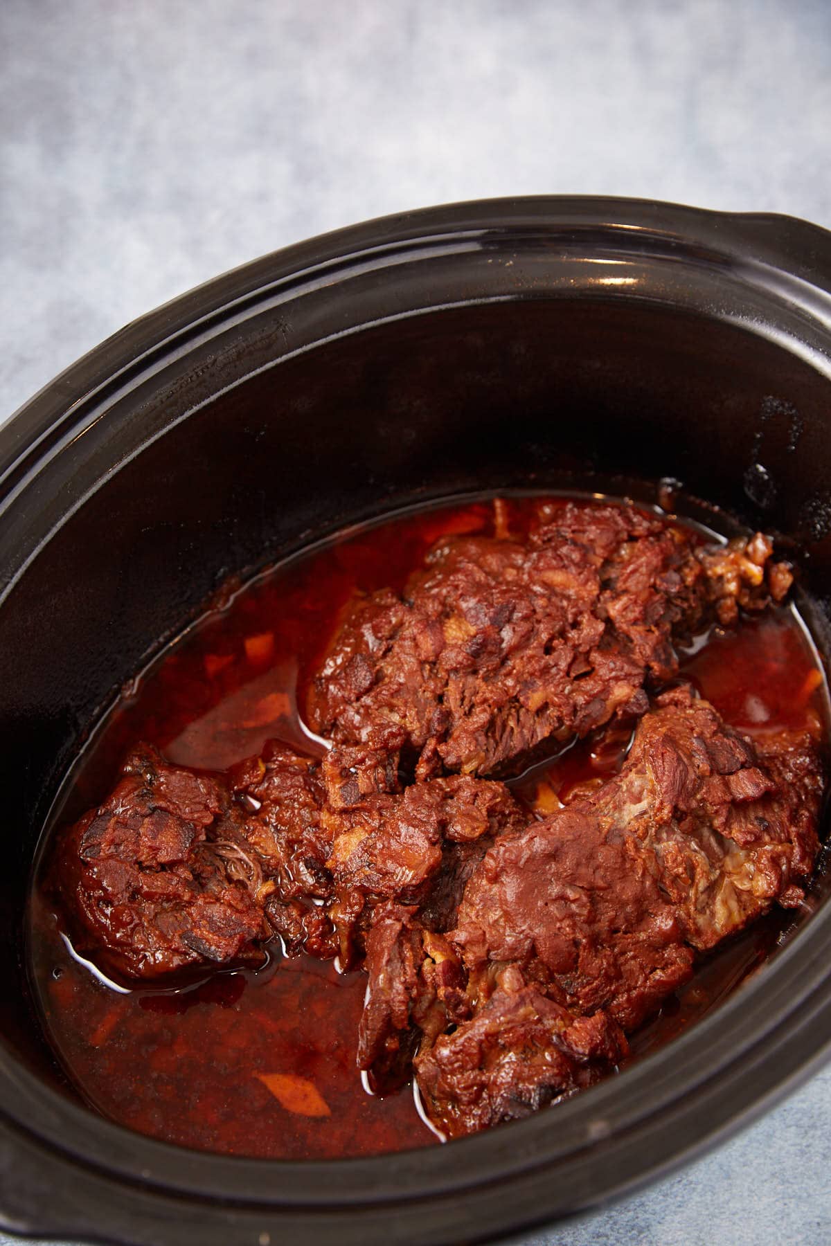 A close-up of cooked beef chuck roast in a slow cooker, sitting in a rich, dark red sauce with visible pieces of onion, against a light blue background.