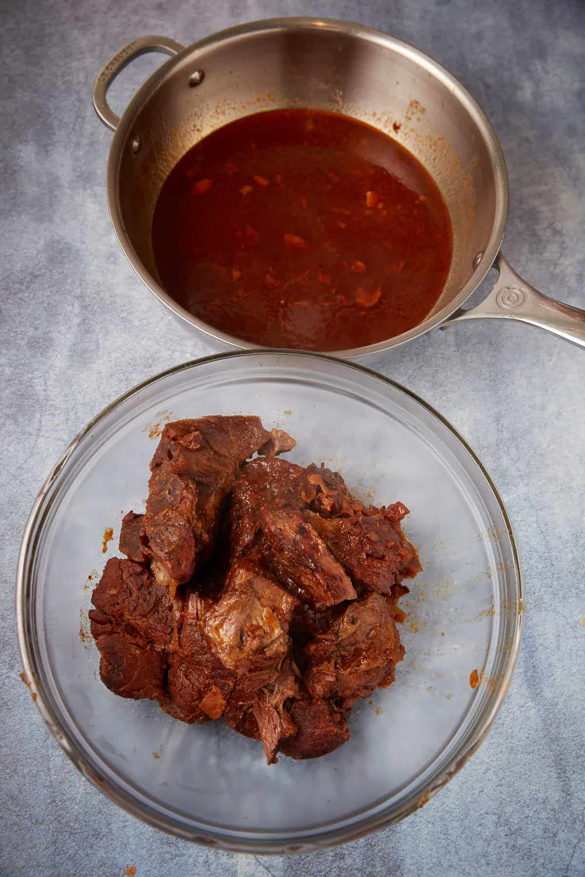 A glass bowl filled with cooked, shredded beef sits in front of a metal pan containing reddish-brown broth with visible oil and small pieces of vegetables. Both are on a gray surface.