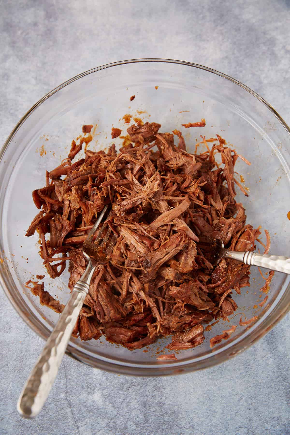 A clear glass bowl filled with shredded cooked beef, with two metal forks resting inside, on a gray textured surface.