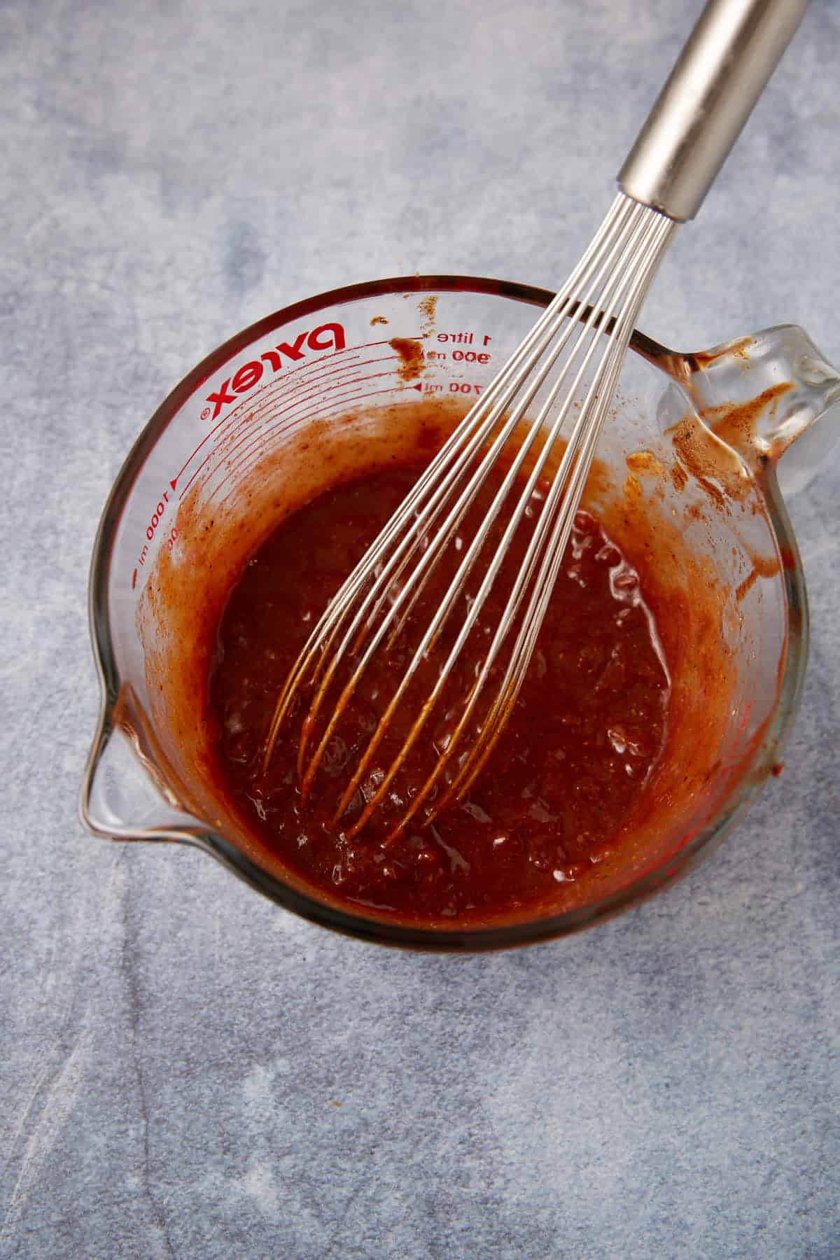 A glass measuring cup with reddish-brown sauce being mixed by a metal whisk, set on a gray textured surface.