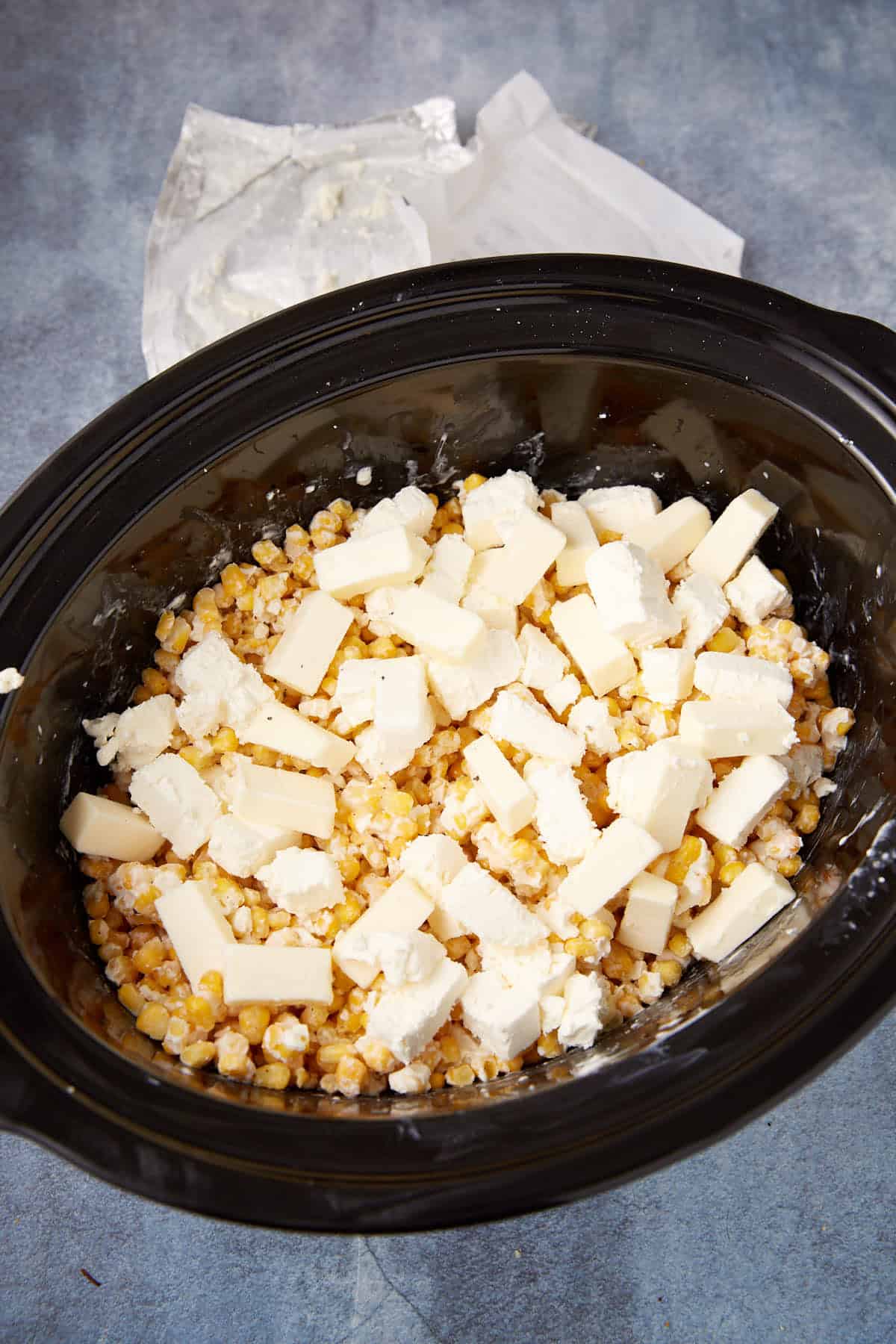 A slow cooker filled with corn kernels, perfect for making slow cooker creamed corn, is topped with chunks of cream cheese and butter and sits on a gray countertop with an unwrapped cream cheese package in the background.