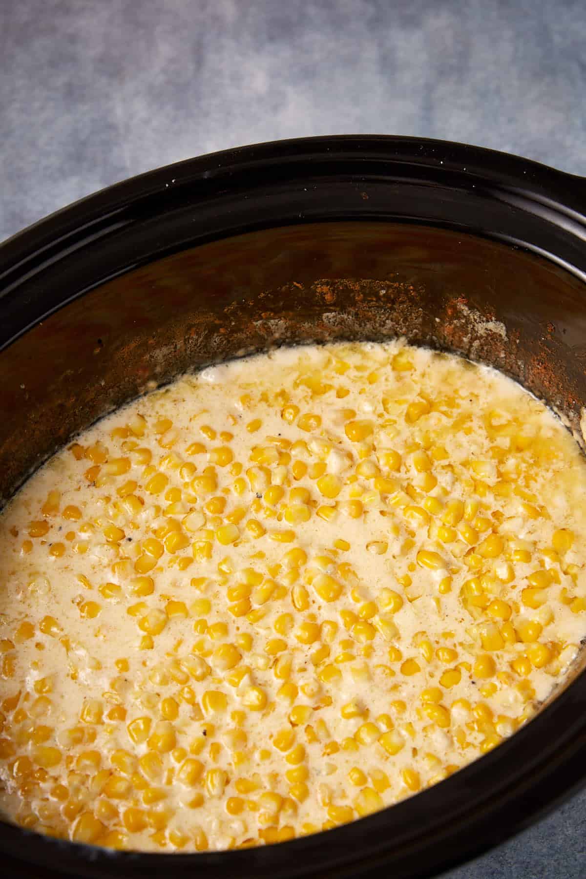 A slow cooker filled with creamy corn, showing yellow corn kernels in a thick, creamy sauce against a blue background.