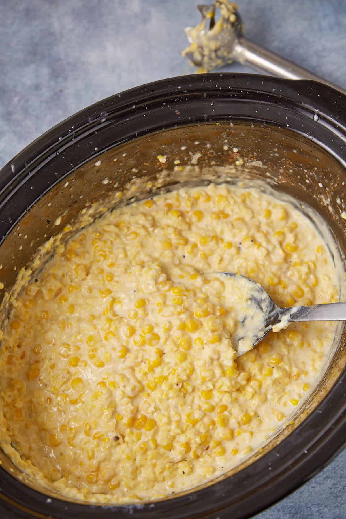 A close-up of slow cooker creamed corn being stirred with a silver spoon. An immersion blender, coated with the creamy mixture, rests nearby on a light gray surface.