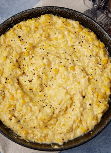 A bowl filled with creamy yellow corn pudding, topped with a sprinkle of black pepper, sits on a cloth napkin with a spoon nearby.