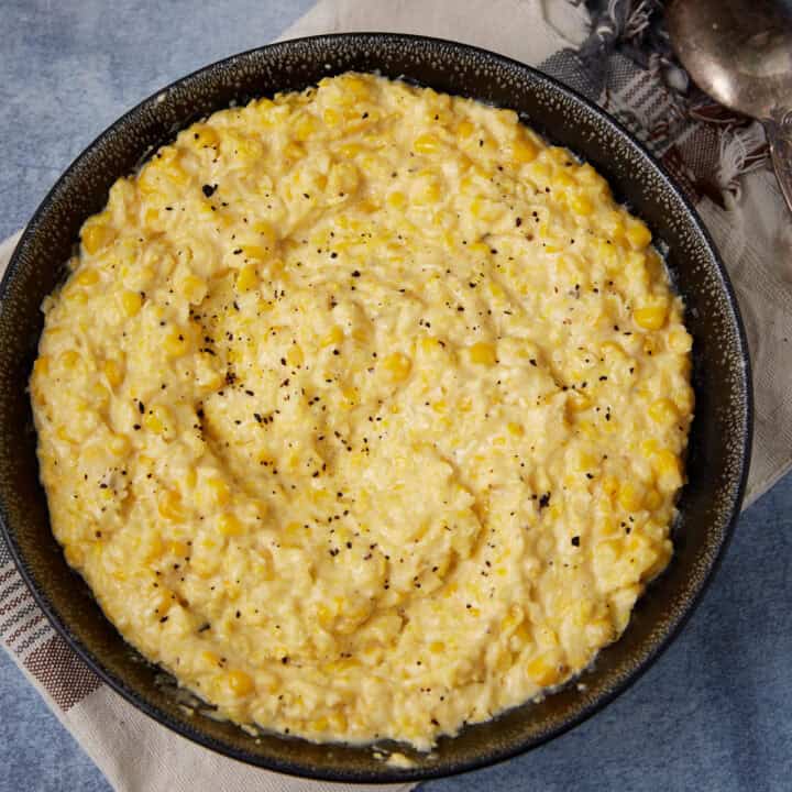 A bowl filled with creamy yellow corn pudding, topped with a sprinkle of black pepper, sits on a cloth napkin with a spoon nearby.