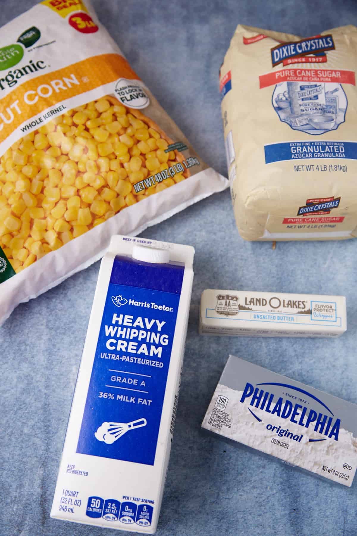 A bag of frozen cut corn, a bag of granulated sugar, a carton of heavy whipping cream, a stick of unsalted butter, and a block of Philadelphia cream cheese on a blue surface.