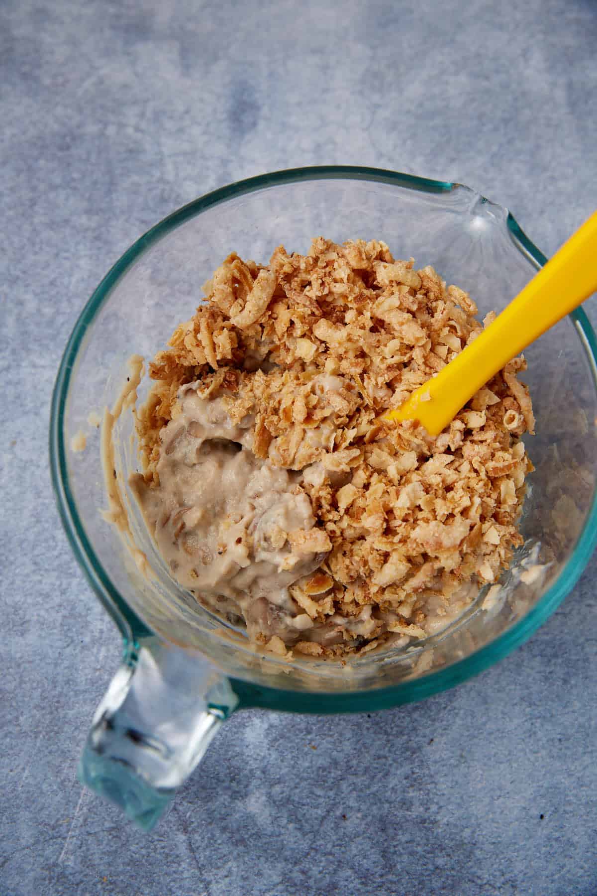 A glass mixing bowl containing a creamy mixture topped with crispy fried onions, being stirred with a yellow spatula, on a gray surface.