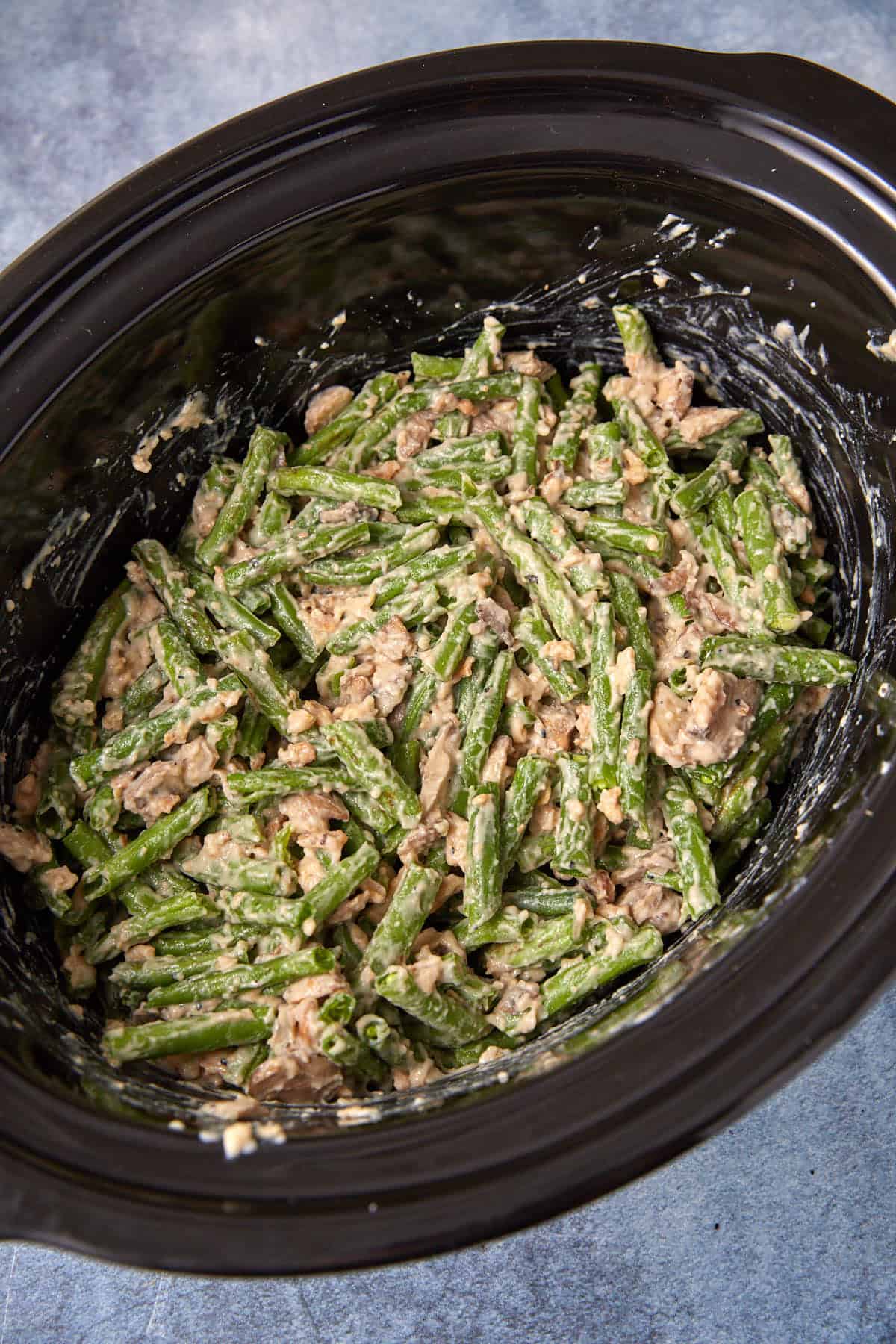 Green bean casserole with creamy sauce and mushrooms inside a black slow cooker, shot from above on a gray surface.