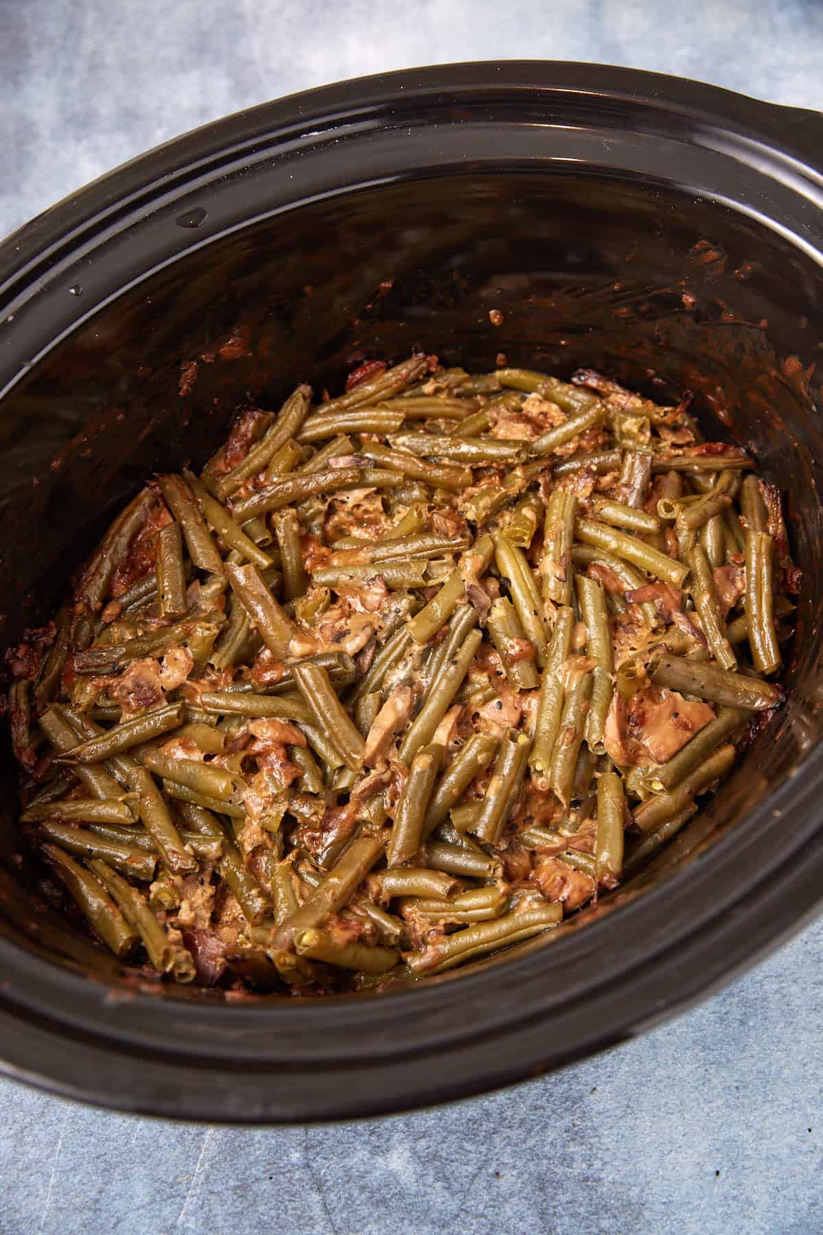 Green beans mixed with pieces of bacon and seasonings are cooked together in a black slow cooker on a light-colored surface.