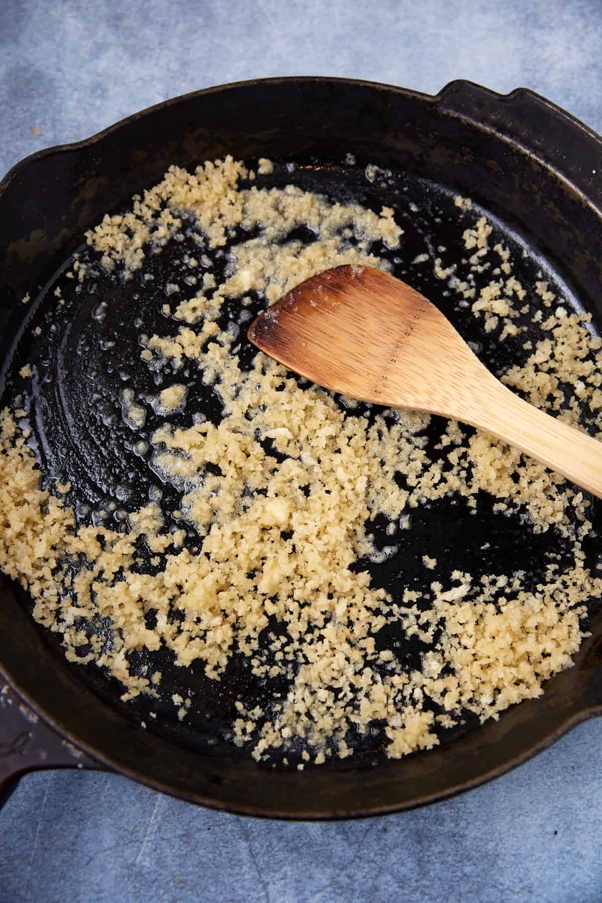A cast iron skillet with browned, crumbled garlic being sautéed in oil, stirred with a wooden spatula on a gray countertop.