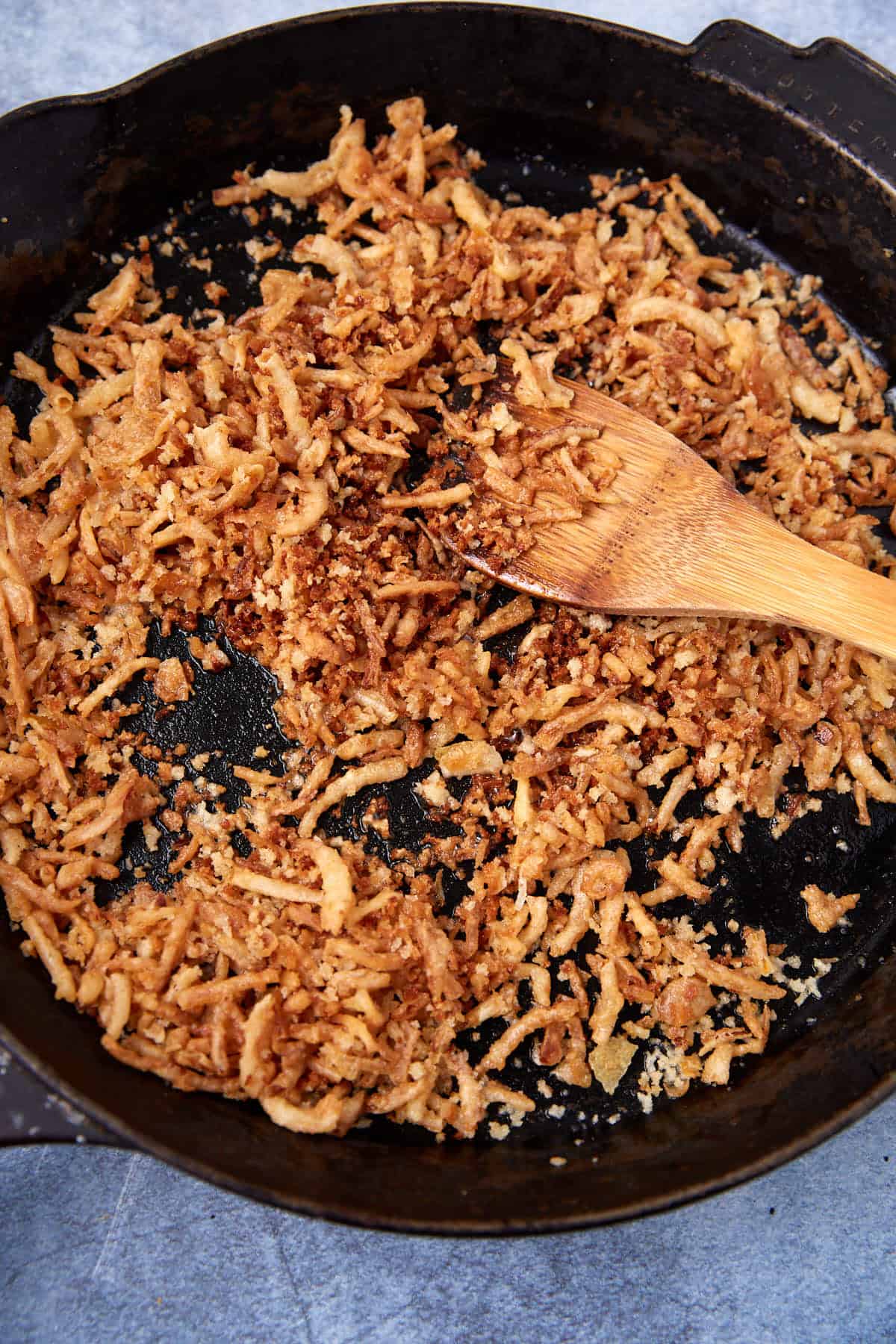 Crispy fried onions scattered in a black cast iron skillet with a wooden spatula resting on the onions; skillet sits on a gray surface.