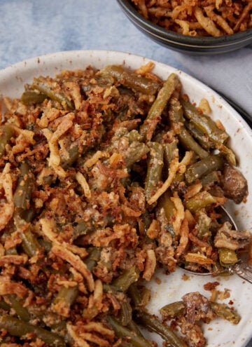 A plate of green bean casserole topped with crispy fried onions, with a spoon on the side. A small bowl with more casserole sits nearby on a light gray cloth.