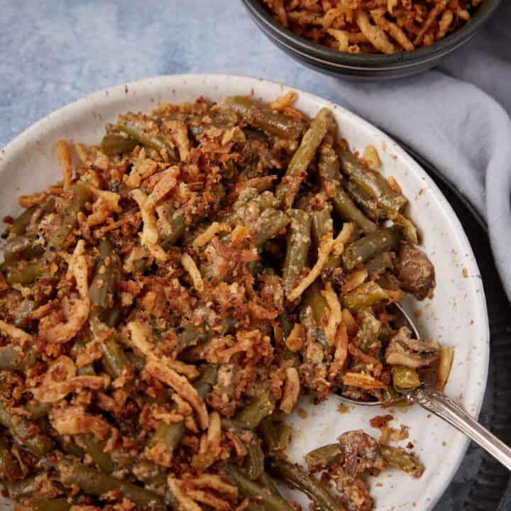 A plate of green bean casserole topped with crispy fried onions, with a spoon on the side. A small bowl with more casserole sits nearby on a light gray cloth.