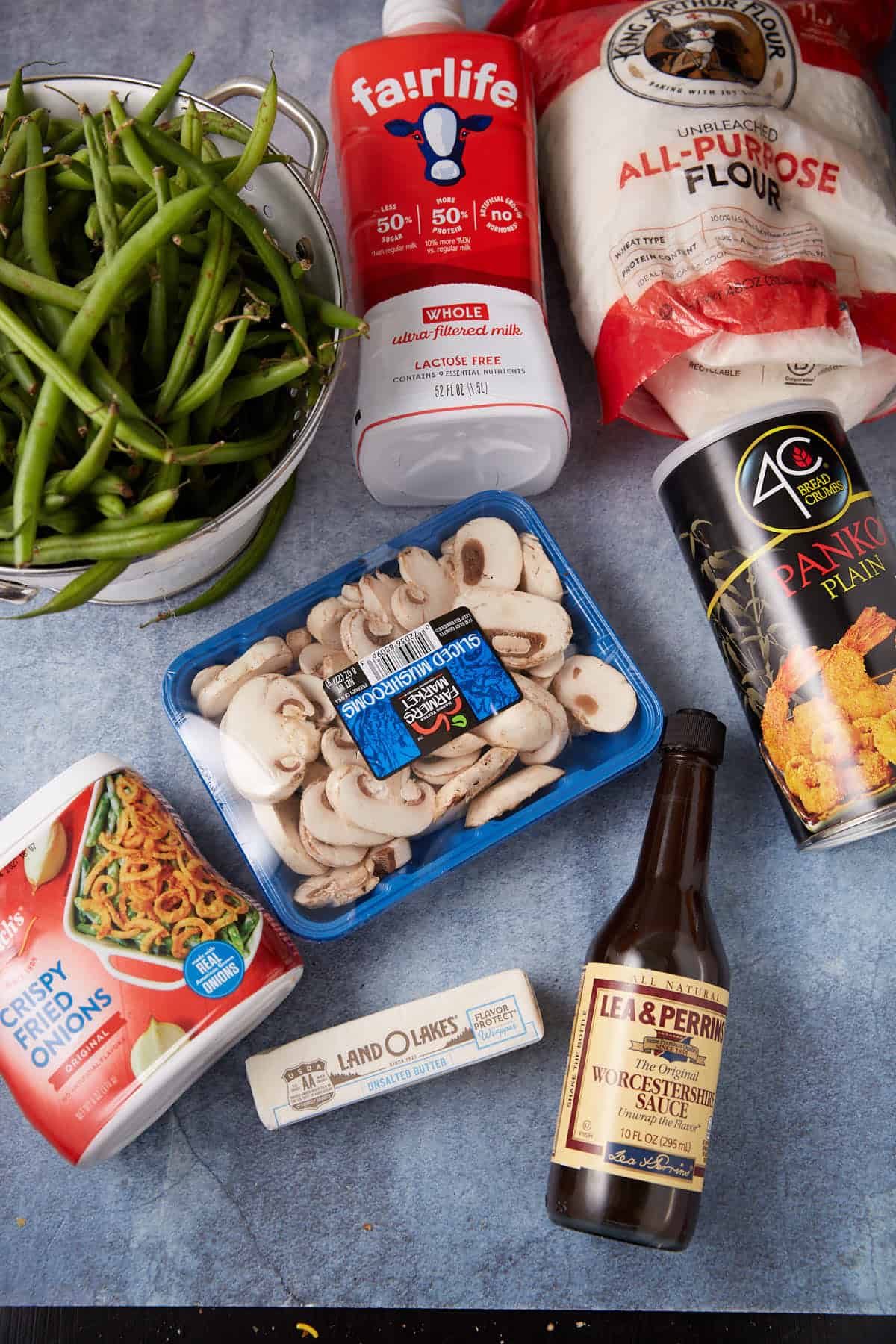 A flat lay of fresh green beans, sliced mushrooms, a stick of butter, milk, all-purpose flour, panko breadcrumbs, crispy fried onions, and a bottle of Worcestershire sauce on a blue surface.