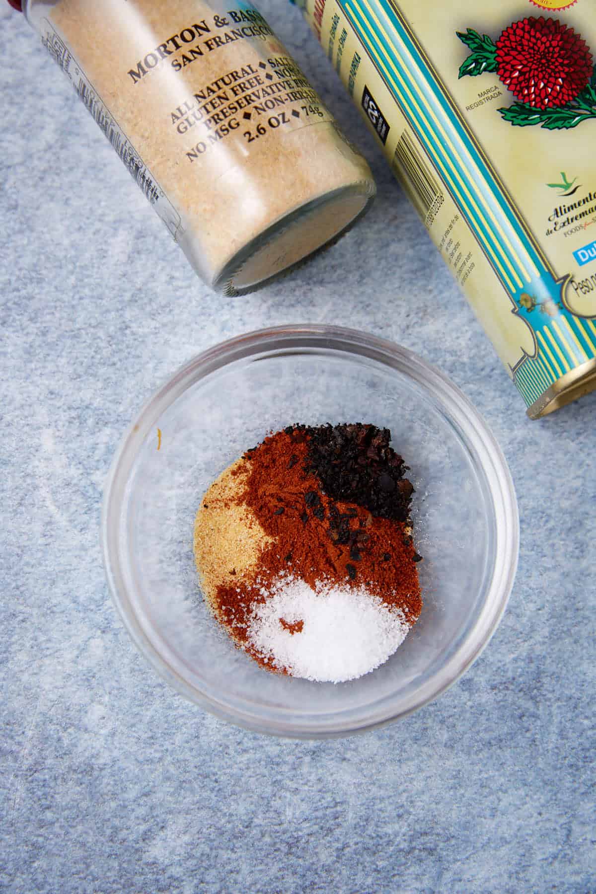 A small glass bowl filled with various spices, including salt, paprika, black pepper, and others, is placed on a blue surface, with containers of seasonings nearby.