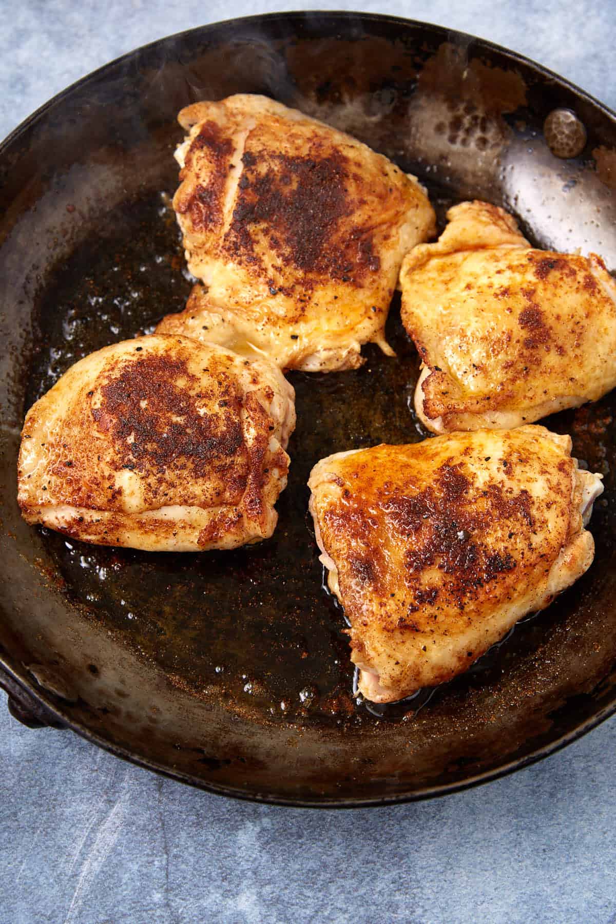 Four browned, seasoned chicken thighs are cooking in a dark, round skillet on a light blue surface. The chicken has crispy, golden skin with visible spices.