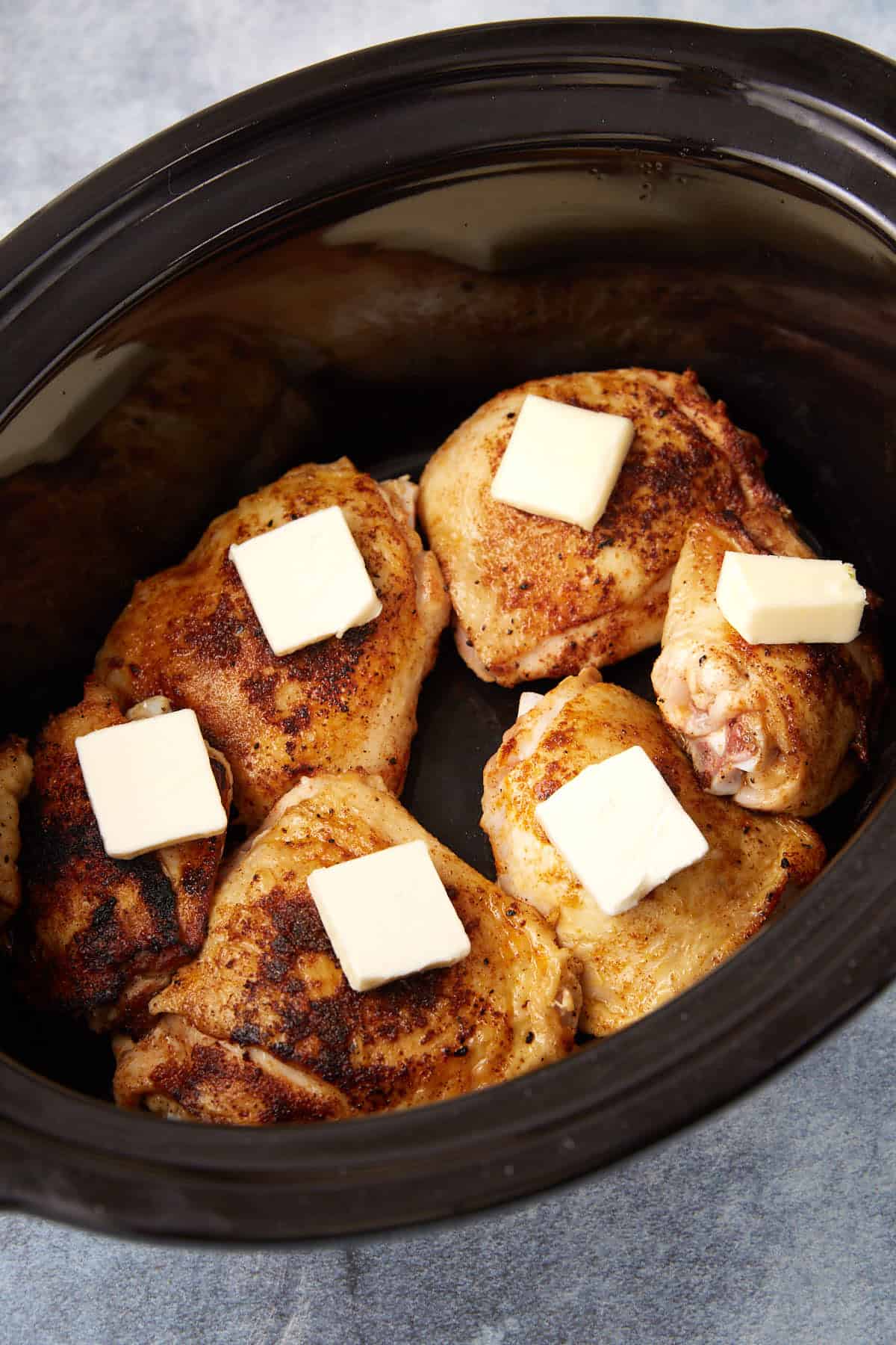 Six browned chicken thighs topped with square pats of butter arranged in a black slow cooker.