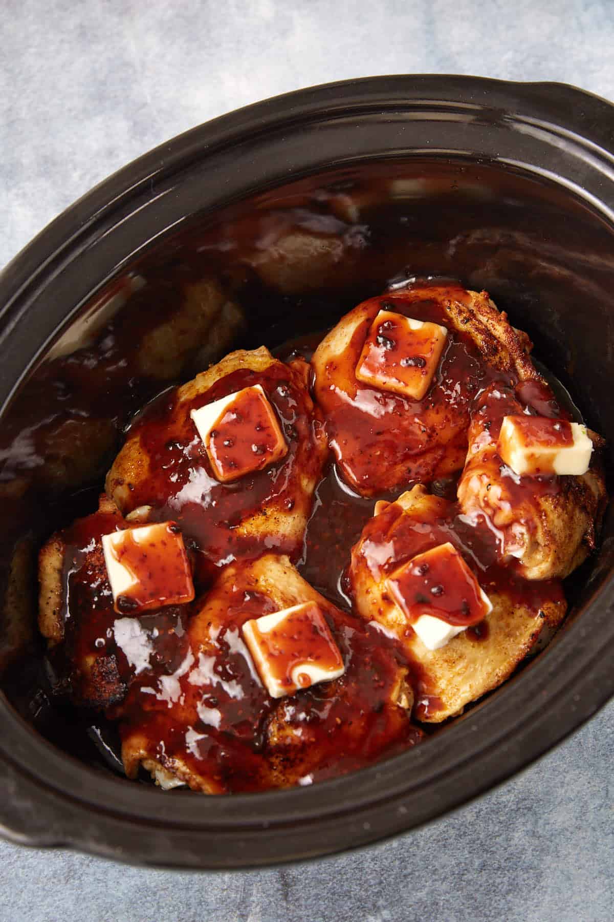 Four pieces of cooked meat topped with dark sauce and butter pats inside a black slow cooker, set against a light blue background.