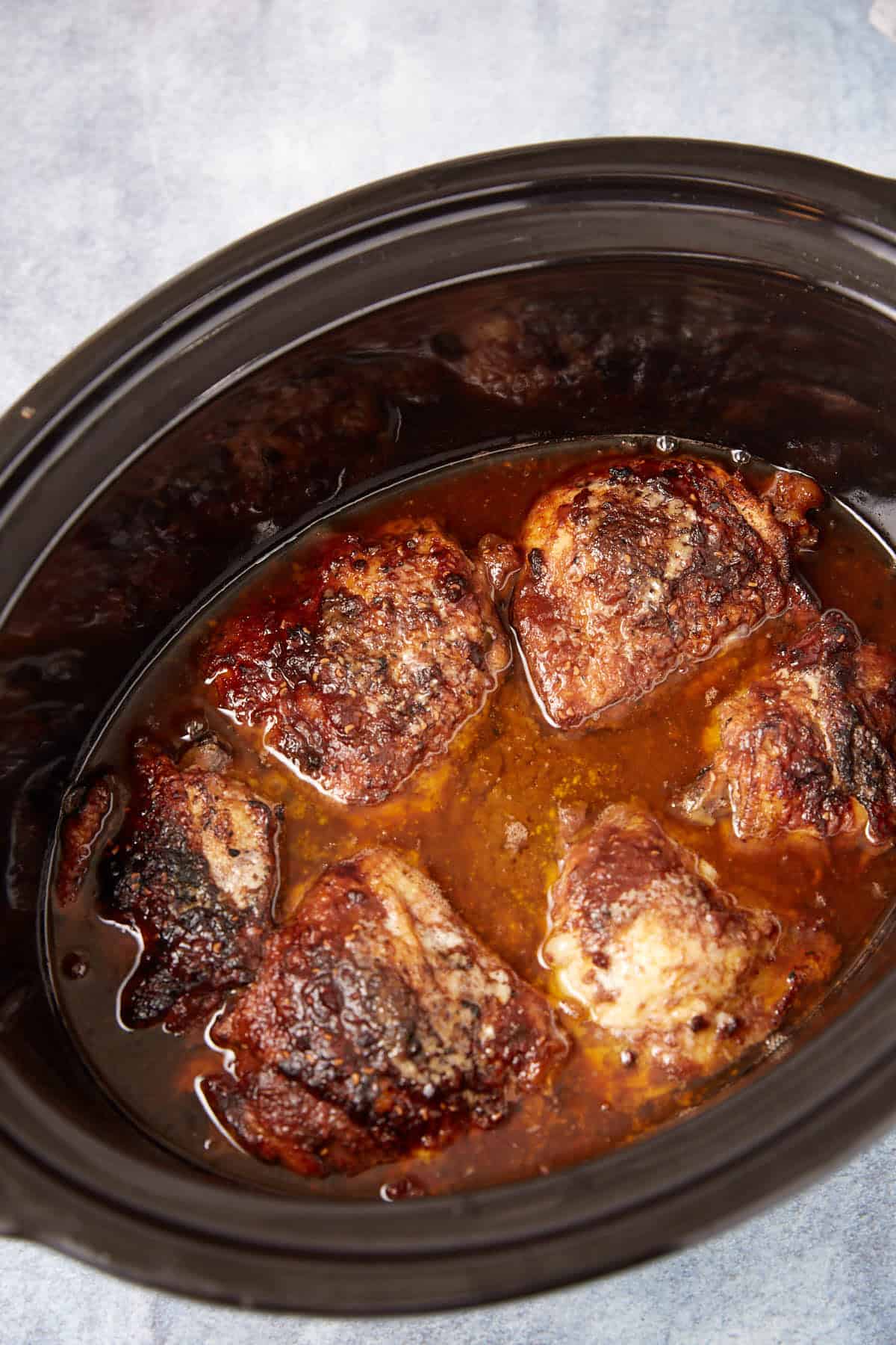 Five browned chicken thighs cooked in a slow cooker, sitting in a pool of flavorful juices, with a dark, caramelized surface and some visible seasonings. The slow cooker rests on a light blue countertop.