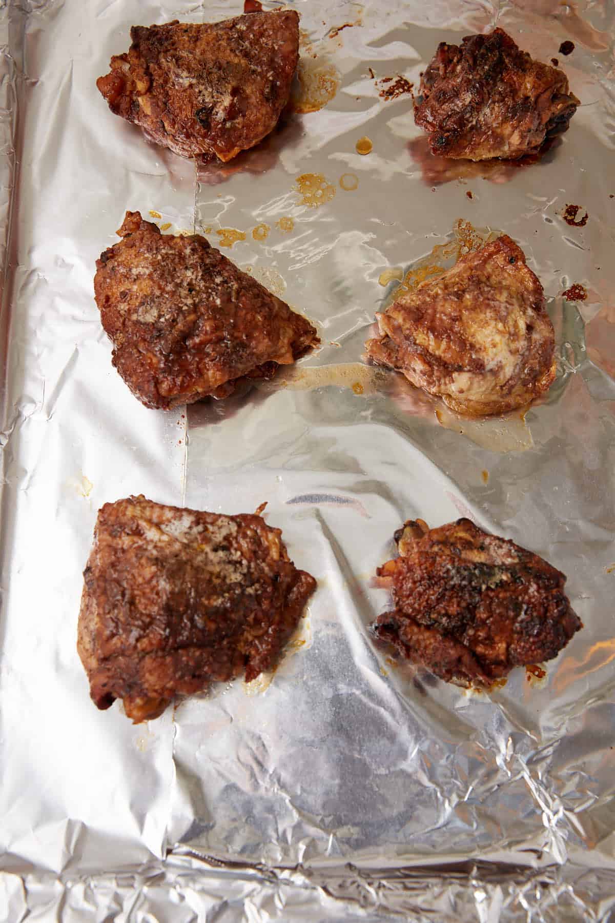 Six browned and cooked pieces of chicken are arranged on a foil-lined baking sheet. The chicken pieces are spaced apart, and the foil shows some spots of oil and juices around them.