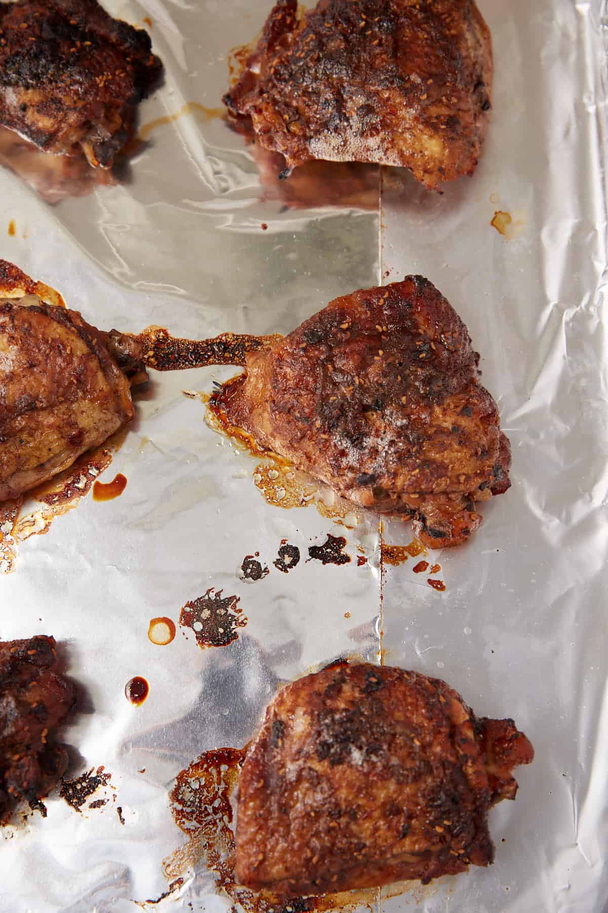 Six pieces of seasoned, roasted chicken thighs on a sheet of crinkled aluminum foil, with some drippings and crispy bits visible around the pieces.